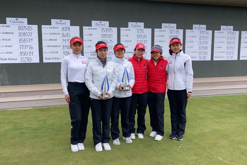 Photo of members of the BU women's golf team. Some wear white zip-ups other where red puffies, and they all wear black pants, white shoes, and red visors. Team's scores are written on large sheets of paper hung up behind them. The six women appear to be Asian or Asian American.