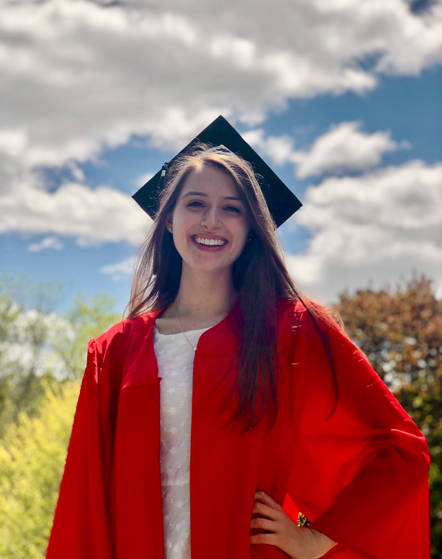 Photo of Leanne Quinn, smiling, in a red graduation gown, and black mortar board. Behind her, white puffy clouds and the tops of trees are seen.