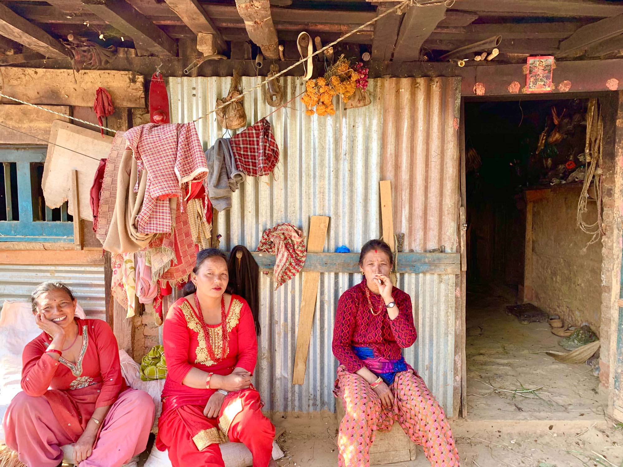 Photo of three Nepalese women wearing red Nepalese garb smiling and sitting in front of a building made of metal and wood scraps. Various clothes hang on a line above their heads.  