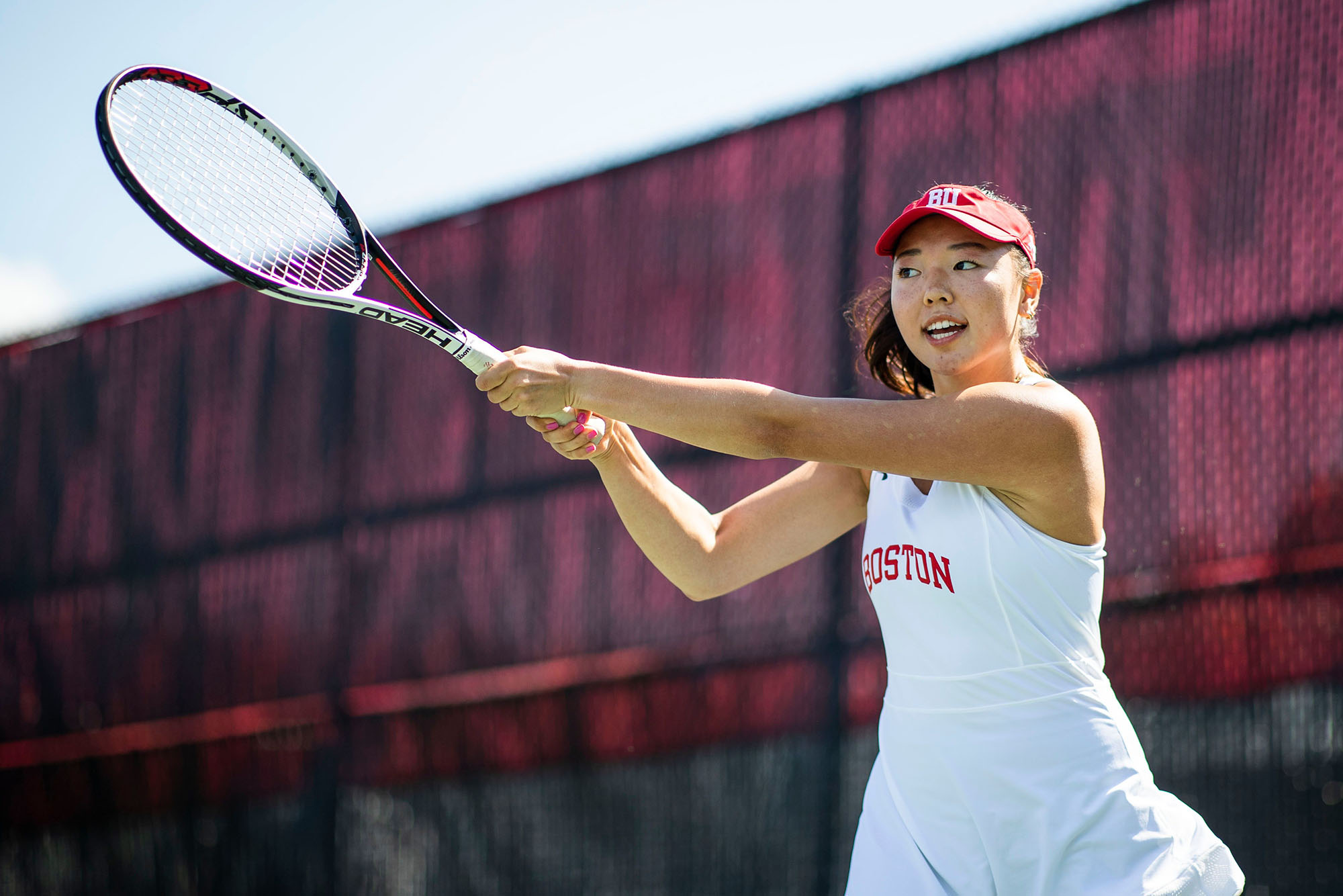 Action photo of BU tennis player Emily Kim. She is wearing a white tennis dress that reads "Boston" in red letters over the chest area and a red BU visor hat. She is captured mid-swing on the tennis court.