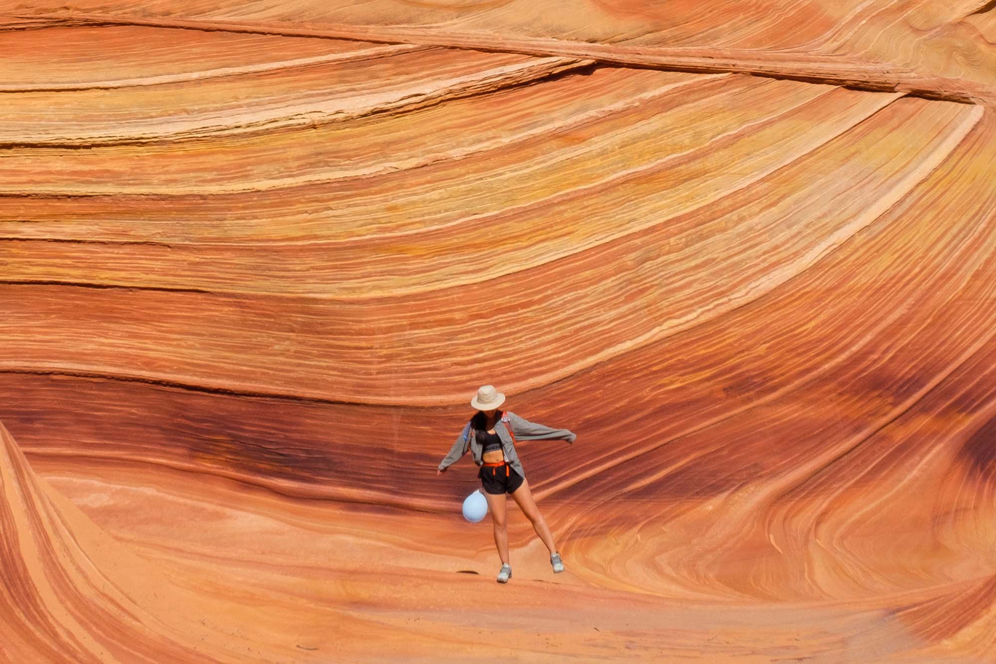 Photo of a person standing on one leg and looking down at their feet as they stand in front of a large orange, striped rock face