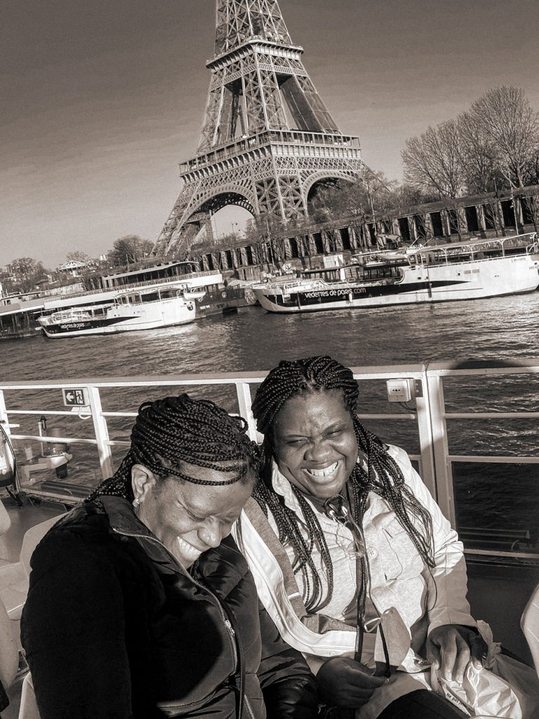 Candid black and white photo of two Black women smiling and laughing on a boat as it travels down a canal. The bottom half of the Eiffel tower can be seen on the shore behind them
