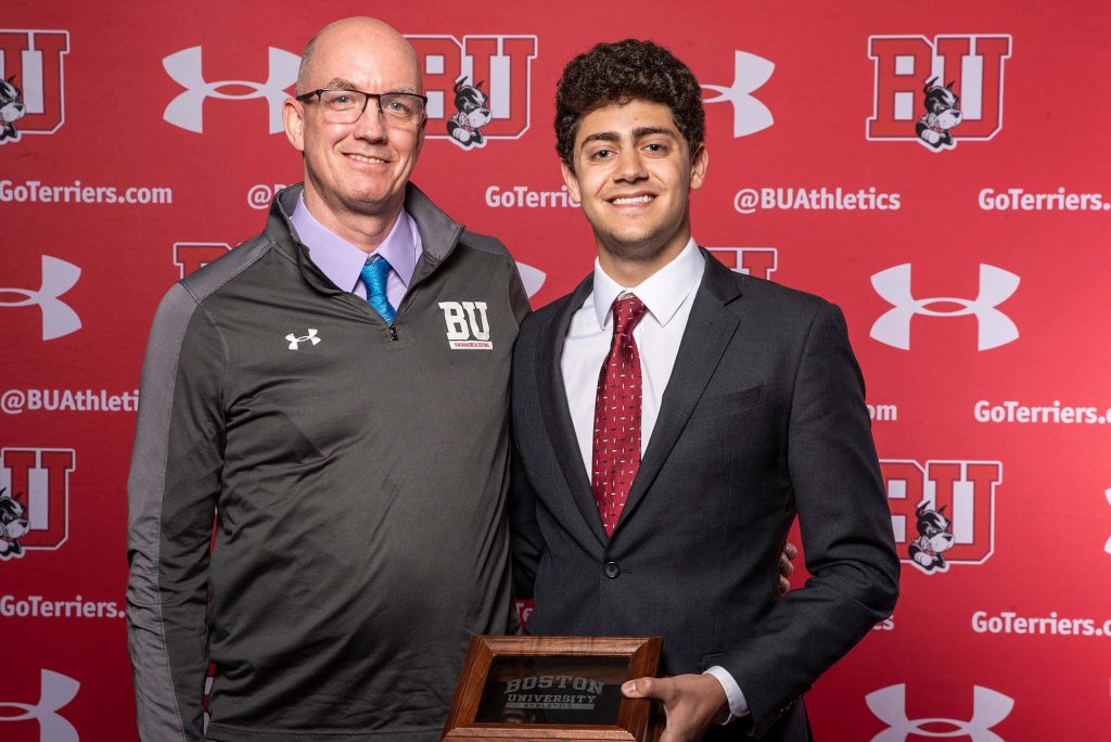 Photo of Michael Nejaime, a young man who wears a suit, red tie, and smiles as he holds his award and poses with Bill Smyth, his coach, who wears a gray BU quarter zip. He is an older white man with glasses.