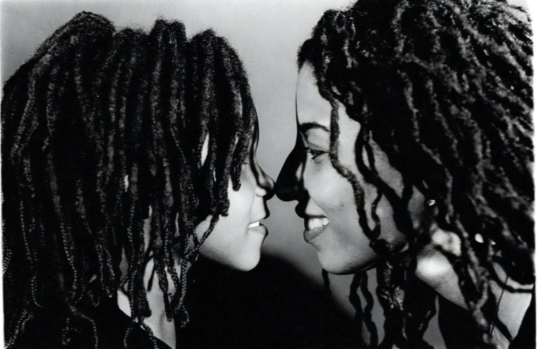 Black and white photo of a Black woman and her child, both with dread locs, standing in front of each other with noses touching, smiling, and looking at each other's eyes.