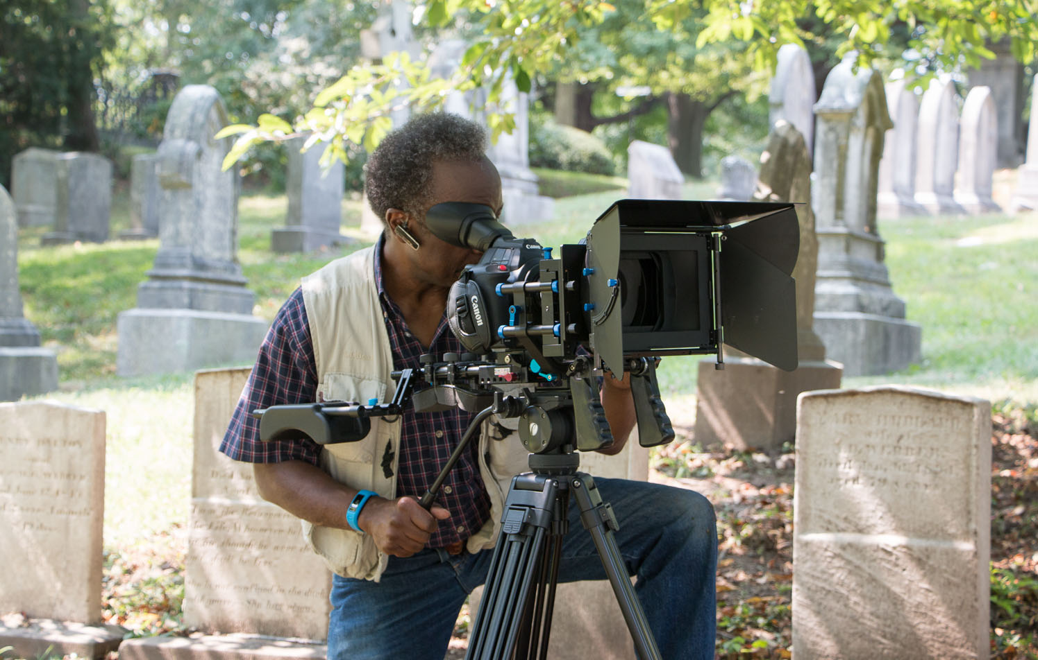 Photo of Roberto Mighty, an older black man with dark grey hair wearing a purple plaid shirt and tan filming vest as he looks through the peephole of a large video camera on set in a cemetery.