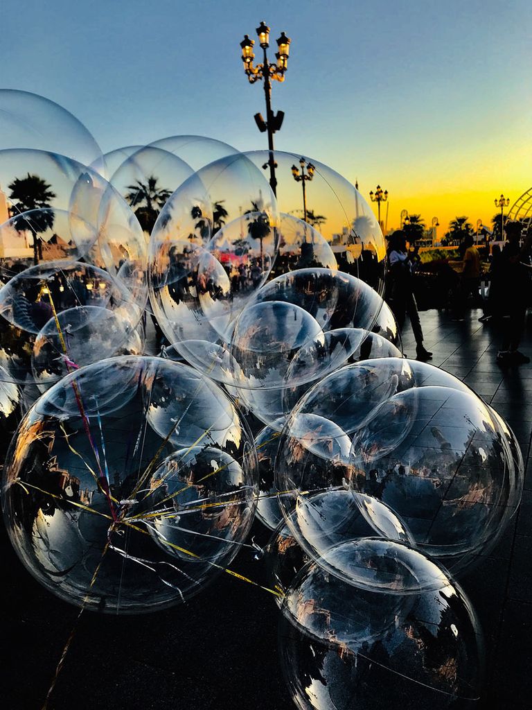 Photo of a large cluster of bubbles being blown in front of a scenic view of a walkway near the end of a sunset. The scene can be seen being reflected in the bubbles.