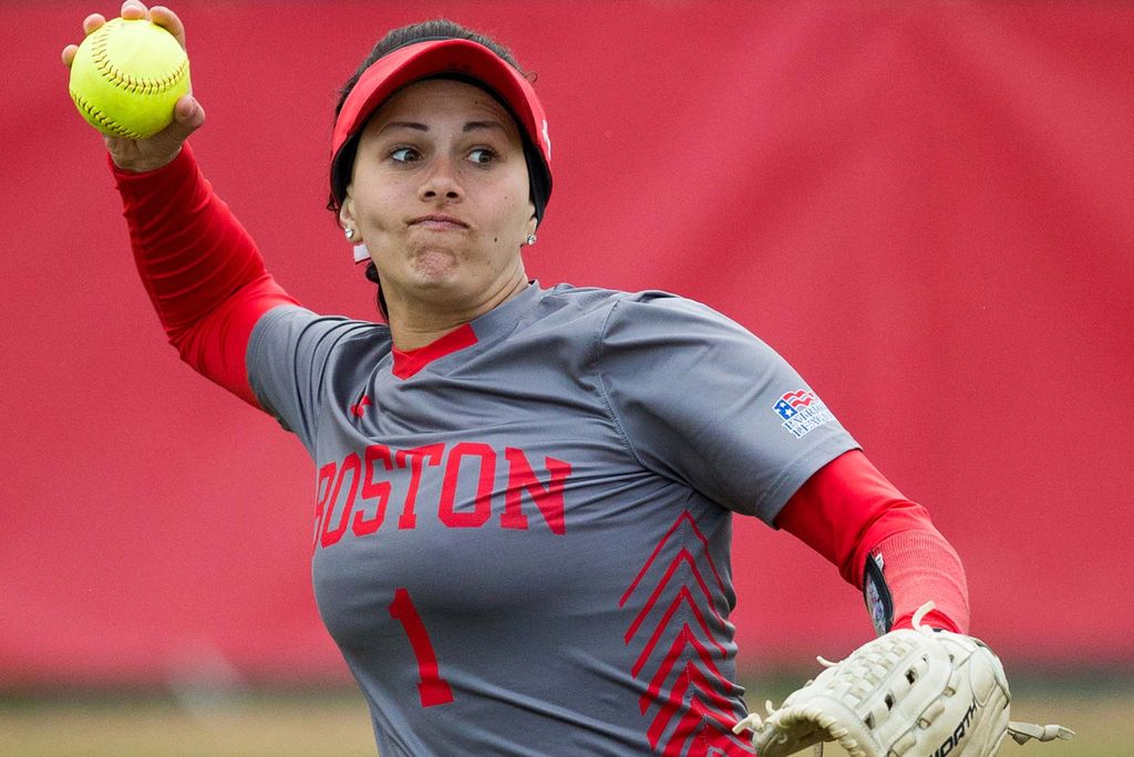 Photo of Marina Sylvestri during a softball game. She wears a gray and red jersey, red visors, and swings her arm back and she goes to the throw the ball. She wears a glove in her left hand.