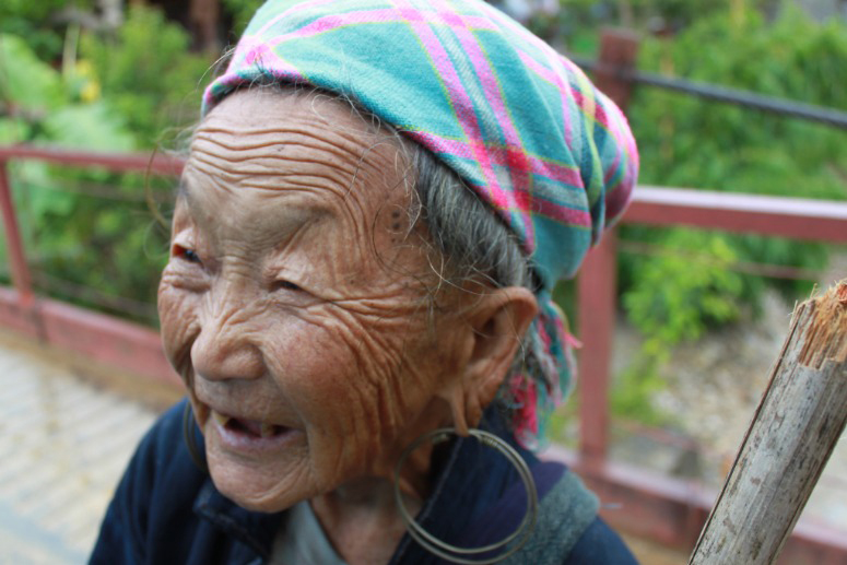 Close up photo of a Vietnamese woman looking and smiling to the left. She wears a light blue and pink striped bandana and large grey hoop earrings that pull at her lobes.