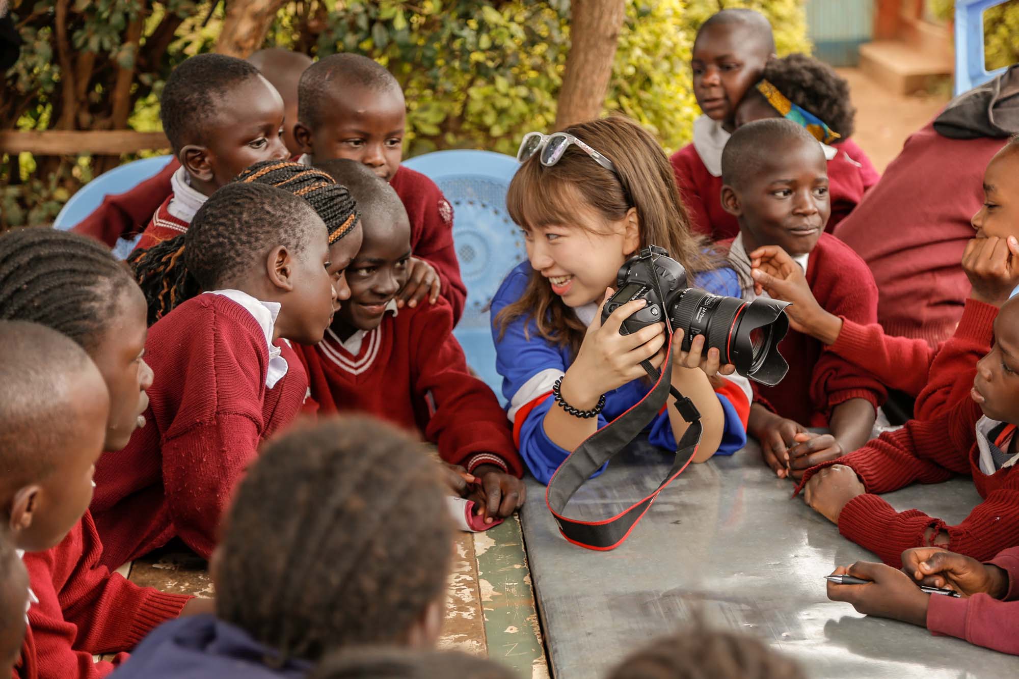 Photo of a young Asian woman with shoulder length brown hair showing her DSLR to a group of Kenyan schoolchildren in red uniforms who are crowded around her.