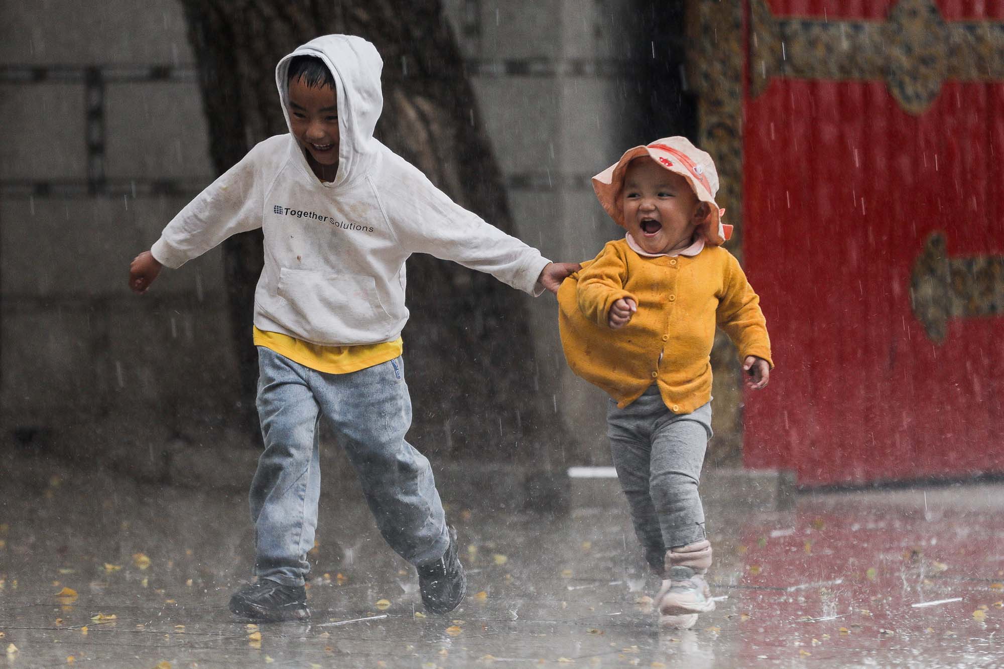 Photo of a Tibetan child having fun with his little sister in the heavy rain. They both are captured mid run as they laugh and play in the rain.