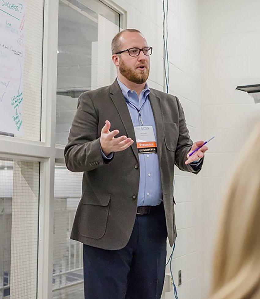 Photo of Jeff Murphy (Questrom’06) in a grat jacket and light blue button down explaining something at the front of a room. He appears to be at a conference as he wears a badge around his neck.