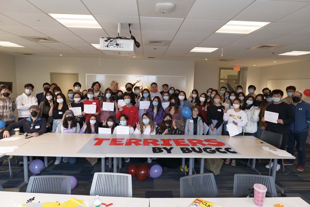 Photo of students in Boston University Global China Connection who created the "Largest LEGO® Brick Word". The lego word is on a table in front of the group and res "Terriers by BUGCC" in black and red on a gray background. The students wear face masks and some hold up paper certificates.