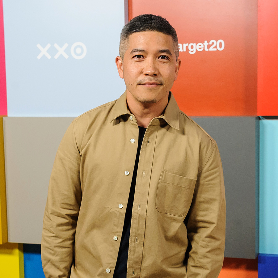 Photo of Thakoon Panichgul standing in front of a colorful light blue, red, and grey backdrop. He wears an open, tan collared shirt, black undershirt, and jeans
