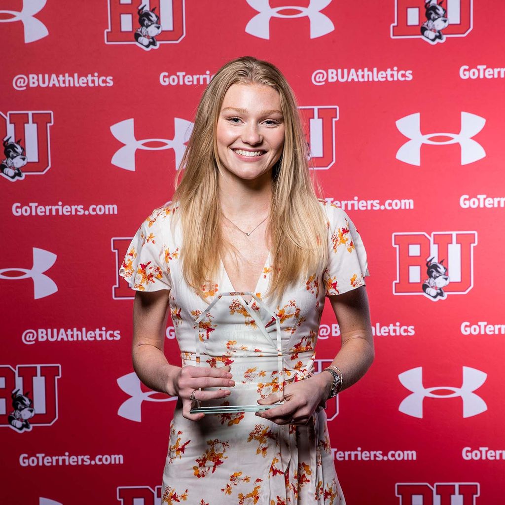 Photo of women's track & field athlete Emma Lawrence. She is shown holding a small, clear trophy in front of her as she wears a white dress with yellow and red florals. She stands in front of a red backdrop with the BU school logo, the Under Armour logo, and the words "@BUAthletics" in an intermittent pattern.