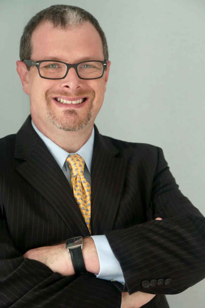 Photo of Scott Singer in a black suit and yellow, blue-spotted tie, with his arms crossed, smiling on a neutral background.