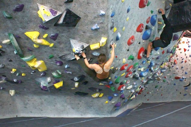 Photo of a young woman in a brown tank top and green pants climbing up a cave wall at Cambridge CRG. Another young person at right in a black sweatshirt is also seen climbing.