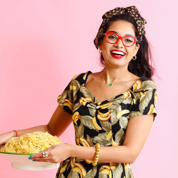 Photo of Chef Priyanka Naik, wearing a dress with a banana and leaf pattern, holding a heaping plate of spaghetti and wearing her signature red glasses. She smiles wide and stands in front of a pink background.