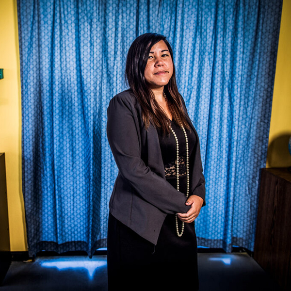 Photo of Erica Mosca posing in a classroom. She wears a dark grey blazer over a long black dress and stands in front of blue curtains