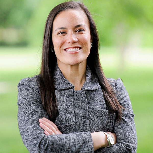 Photo of Gina Ortiz in a gray blazer with her arms crossed, smiling, with green space blurred behind her.