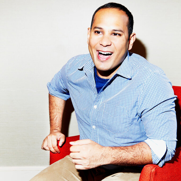 A portrait photo of Sopan Deb laughing. He sits in a red armchair and wears a light blue collared shirt over a dark blue shirt