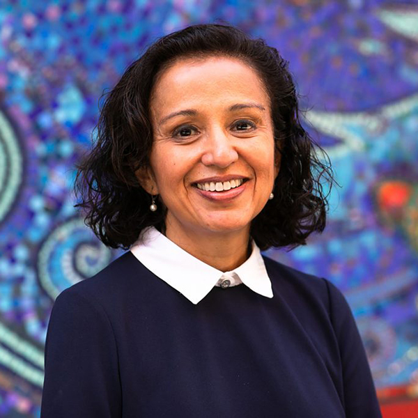 photo of Manjusha Kulkarni, an Indian woman, wearing a blue sweater over a white collared shirt. She stands in front of a bright blue mosaic mural.