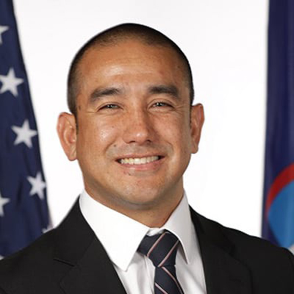 Headshot of BU alum and attorney general of Guam, Leevin Camacho. A tan man wearing a black suit and tie, poses and smiles for the camera in front of the Guam and American flags.