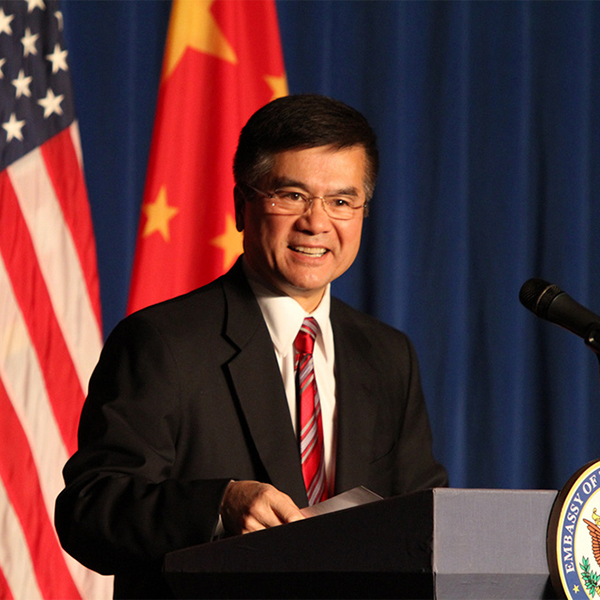Gary Locke in Beijing while serving as US Ambassador to China. He smiles and stands at a wooden podium. Both the American and Chinese flags stand behind him.