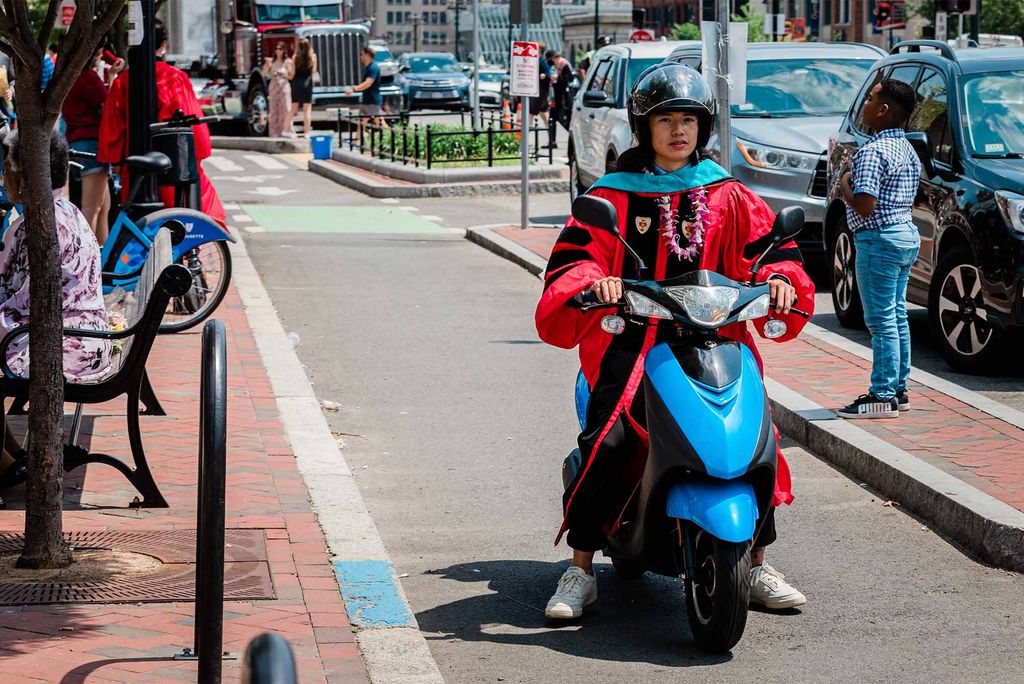 A student wearing a red robe with black stripe and a black sash, and purple flower lei around their neck, rides down Comm Ave in the bike lane on a bright blue scooter. other students processing can be seen in the background.