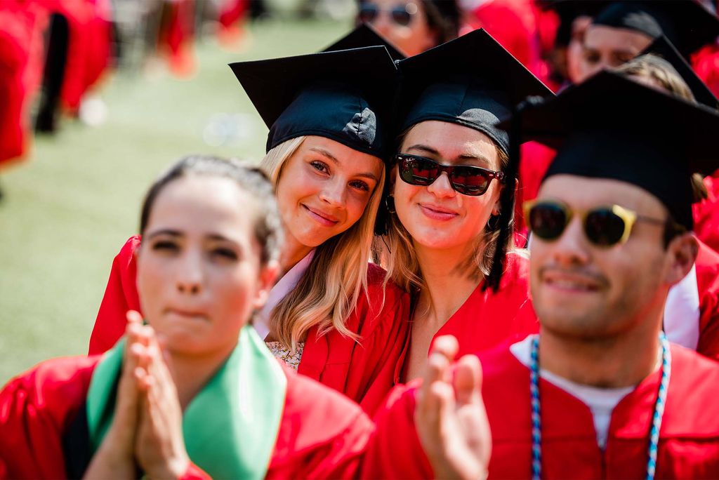 In focus are two young White woman who smile in the red robes and black mortarboards on Nickerson field. Blurred in the foreground is a young woman who wears a green sash and red robe, and a young man with sunglasses and blue beads on.