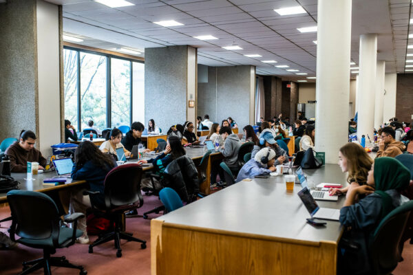 Photo: Students studying at Mugar Memorial Library. They sit at long tables and private spaces as they read and study off books and laptops. A large window brightens the room.