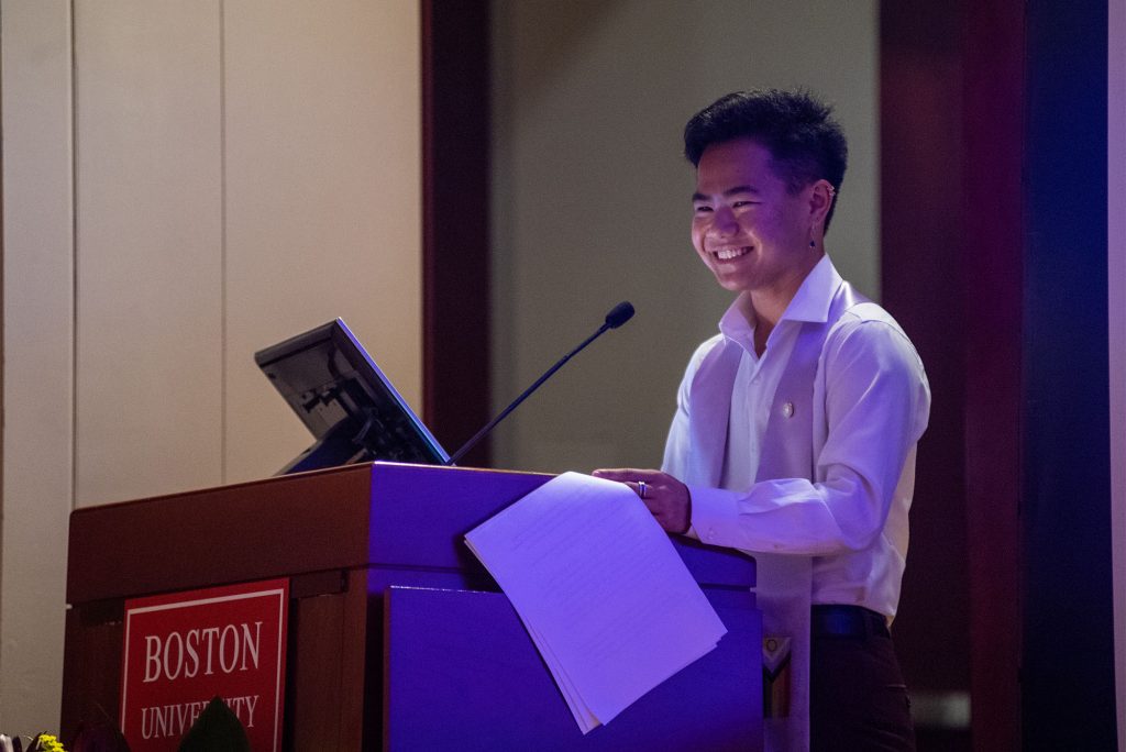 Photo of co-chair Evan Gilbert, an AAPI person, who wears a white button down shirt, has short black hair, and smiles. Gilbert holds a piece of paper and looks towards the audience.