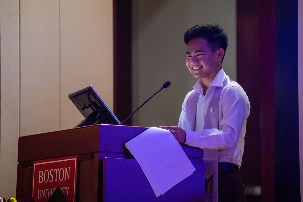 Photo of co-chair Evan Gilbert, an AAPI person, who wears a white button down shirt, has short black hair, and smiles. Gilbert holds a piece of paper and looks towards the audience.