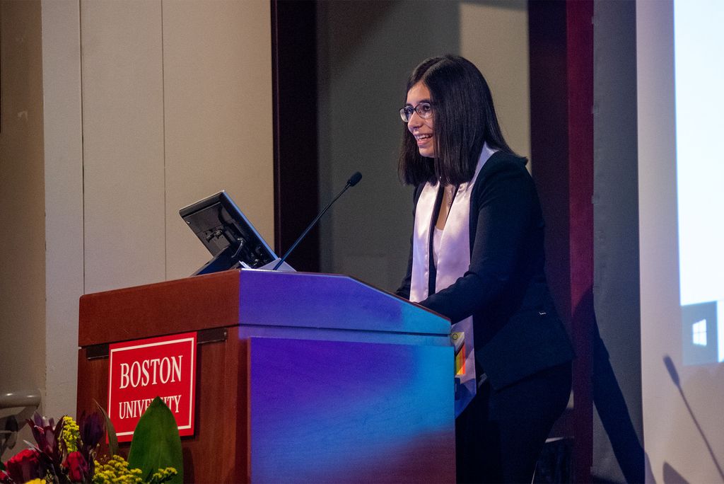 Photo of Christa Rose (CAS'22), Q’s president, who has shoulder length black hair, wears a black sweater, and white sash. Rose stands at the wooden, Boston University podium and smiles as she speak.