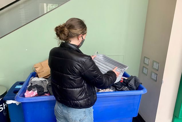 Photo: A masked student places a grey woven basket into a large plastic blue bin filled with other assorted donated items.