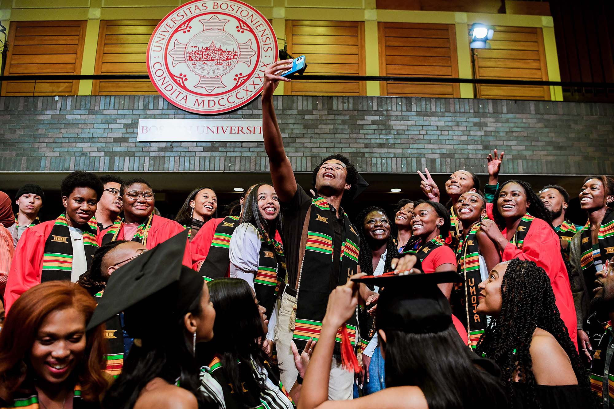 Photo of Ethan Strauther taking a selfie with his friends during the end of the UMOJA induction ceremony. A young black man holds up a cellphone and a large group of Black students crowd around him to be a part of the photo. All wear red graduation gowns and black sashes with Kente stripes. A few students also wear black caps.