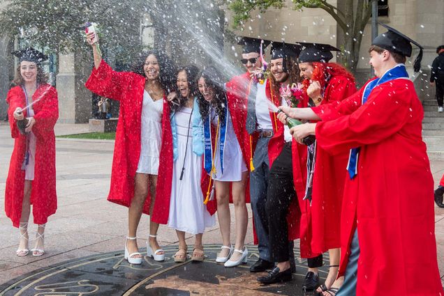 Photo of Charlotte Beatty, from left, Shaina Evans, Jessica Zheng, Kelsey Brown, Conor Kelley, Pedro Henrique De Barcellos Junqueira, Mira Elzanaty, and Sebastian Porreca on the seal in Marsh Plaza. All where red gowns and black caps as they shout in joy as Evans and Porreca shower the group in freshly popped champagne.