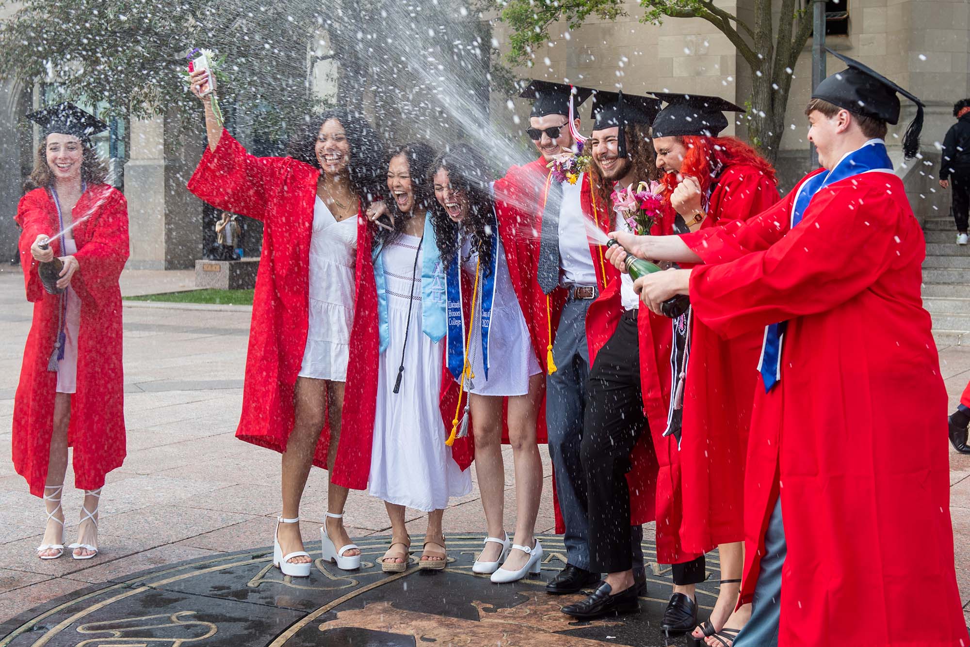 Photo of Charlotte Beatty, from left, Shaina Evans, Jessica Zheng, Kelsey Brown, Conor Kelley, Pedro Henrique De Barcellos Junqueira, Mira Elzanaty, and Sebastian Porreca on the seal in Marsh Plaza. All where red gowns and black caps as they shout in joy as Evans and Porreca shower the group in freshly popped champagne.