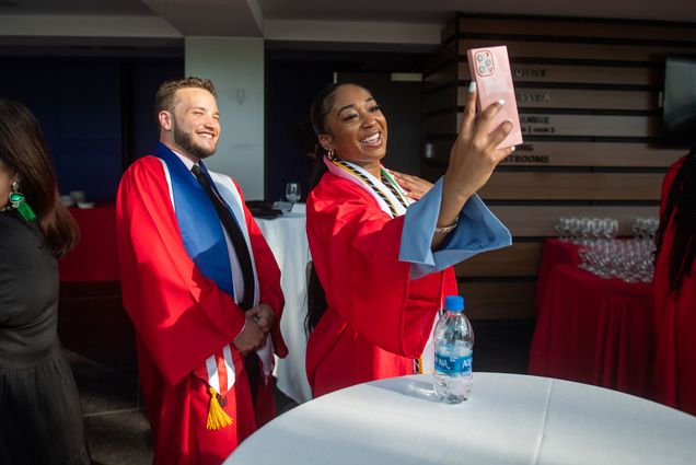 Photo of Oscar Zaysas, left, and Kelsey Russell, at the May 20 BU Posse Graduation Ceremony at Booth Theater. Russel holds up a pink phone as she and Zaysas take a selfie together. Both wear red graduation gowns and blue sashes as they smile towards her phone.