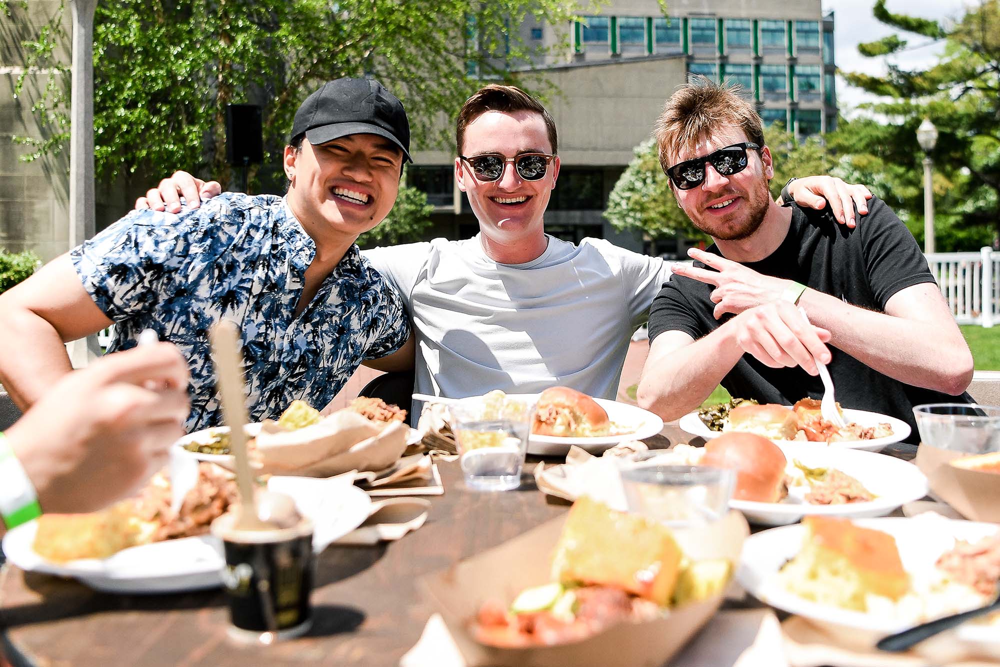 Photo of 3 young men sitting at a table and posing for a photo together. They all wear t shirts and smile together. The man in the middle has his arms around the guys on either side of him. The two on the right wear sunglasses and the one on the far right holds up a peace sign.