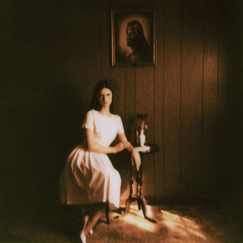 Album cover for Ethel Cain's "Preacher’s Daughter". Cover is an image of Ethel, a young white woman with waist length brown hair, sitting in a chair in an empty room. She wears a white dress and leans casually in the chair in a moody toned room with wooden walls and a photo of what appears to be Jesus framed above her.