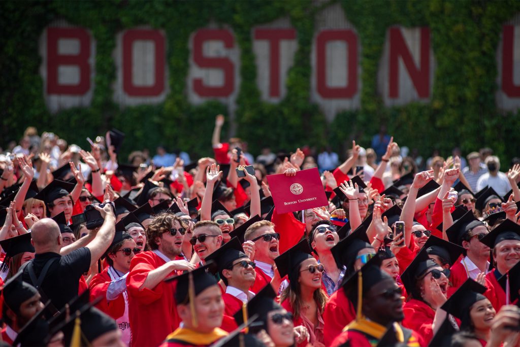 Photo of a large crowd of 2022 BU graduates in red graduation gowns and black caps cheering and standing as they raise their arms towards the stage. One person can be seen holding up a large folder that has the BU Seal on the front and reads "Boston University". A blurry, large "Boston" can be seen on the wall in red in the distance. 