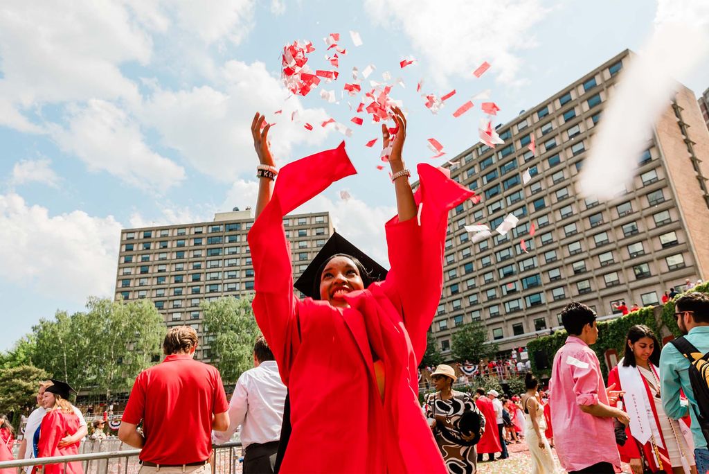 Photo of a young, brown-skinned woman throwing up a handful of red and white confetti into the air. She wears a red graduation gown and black cap as various people mill and talk behind her on the field.