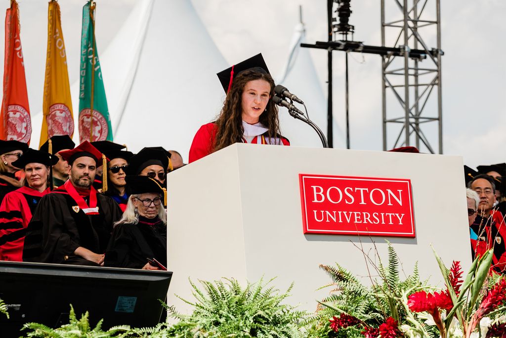 Photo of student speaker Amanda Berke, a white woman with very long brown wavy hair, speaking at a large Boston University podium on stage on nickerson field.
