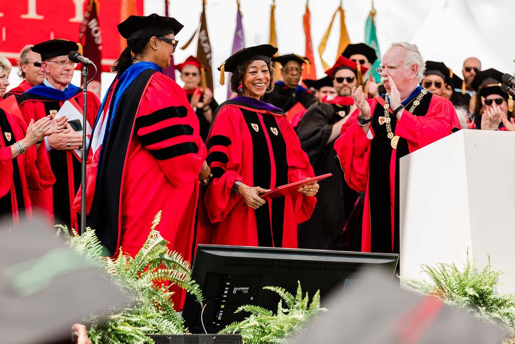 Photo of Gay J. McDougall as she receives her honorary degree. She smiles and holds a red folder in her hands. She is a middle-aged Black woman who wears a red graduation robe with black stripes, black sash, and black hat. At right, Bob Brown claps at a podium. At left, a Black woman in a black hat and purple sash is seen.