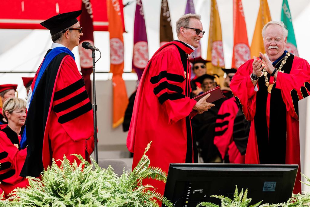 Photo of presumably Richard Shipley receiving his honorary degree on stage. Shipley, seen center, is a white with short gray hair who wears a red robe with black stripes and black sunglasses. At right, Bob Brown claps at a podium. At left, a man in a black hate and purple sash is seen.