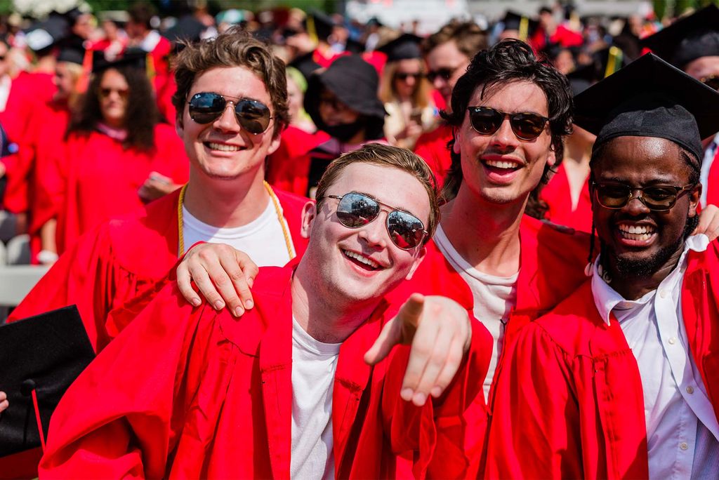 Photo of a group of young men as they process towards Nickerson. They all wear sunglasses; three students are White, one student is Black. They smile wide and wear their red robes unzipped due to the heat.