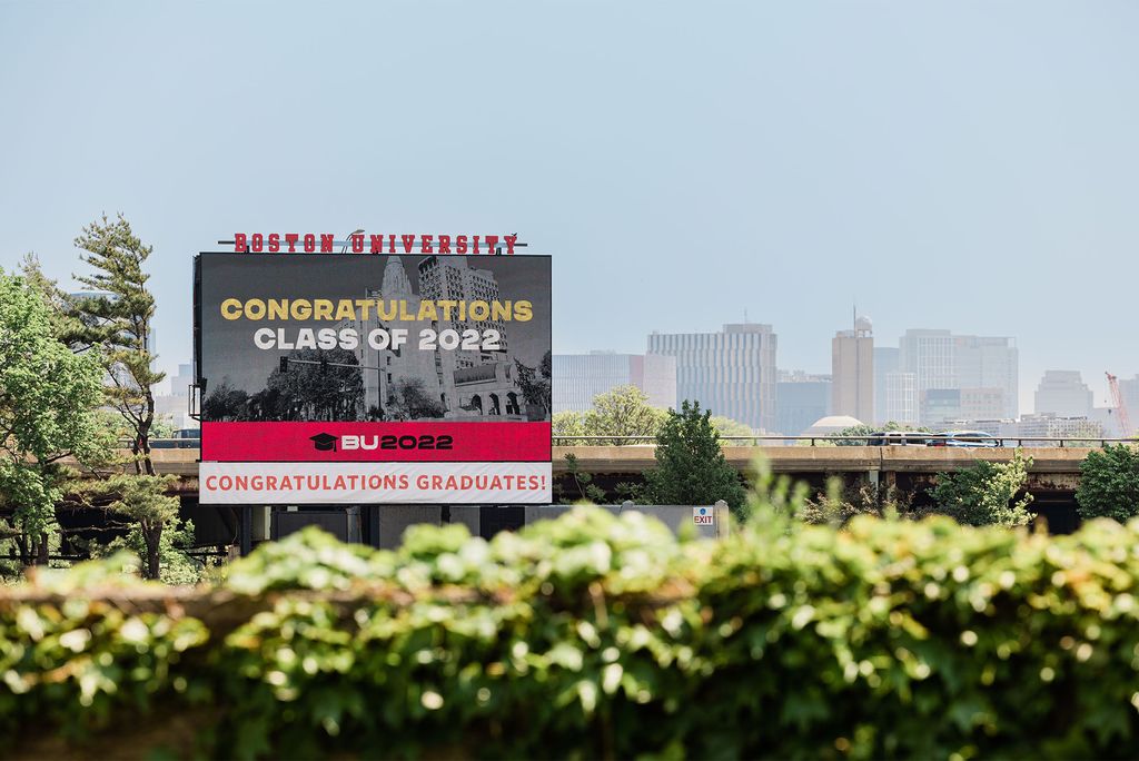 Photo of the Jumbotron at Nickerson field, which reads: Boston University at top. On screen: a photo of Marsh Plaza in Black and White with "Congratulations Class of 2022, BU 2022" overlaid. At bottom, sign reads in red and white: "Congratulations graduates". Hedges are seen in the foreground and the sky is bright blue behind the sign.