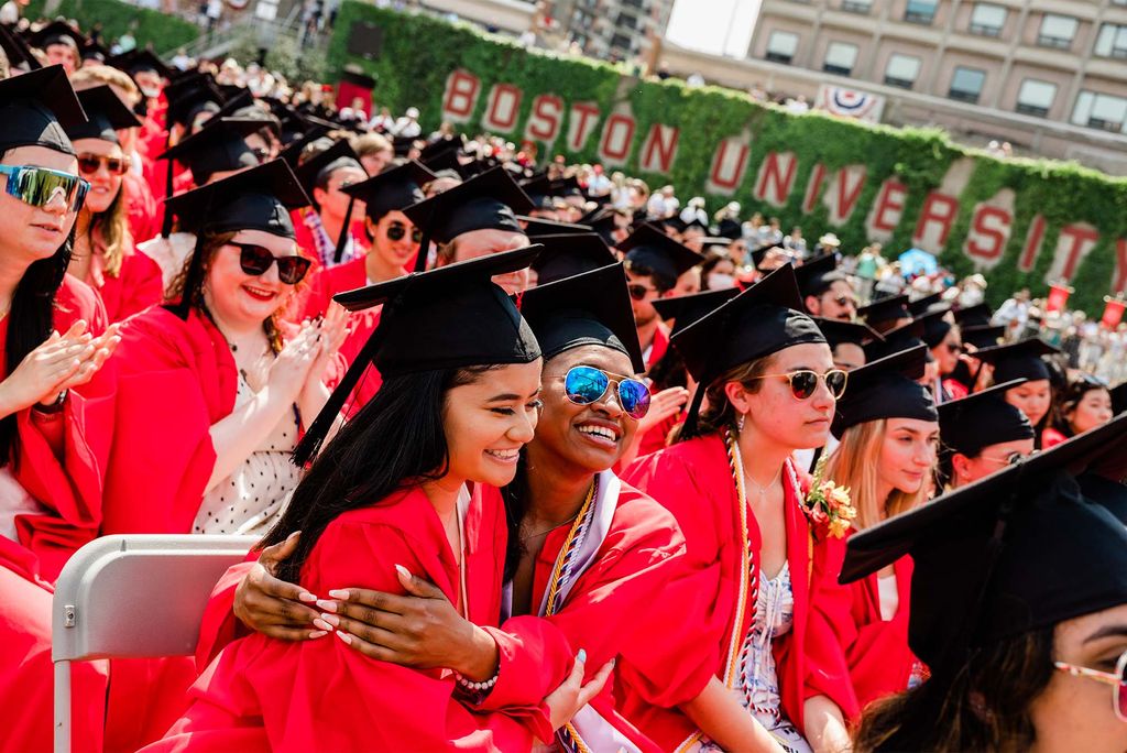 Two students of color in red robes and black mortarboards embrace during commencement. other students and Boston University sign are seen behind them.