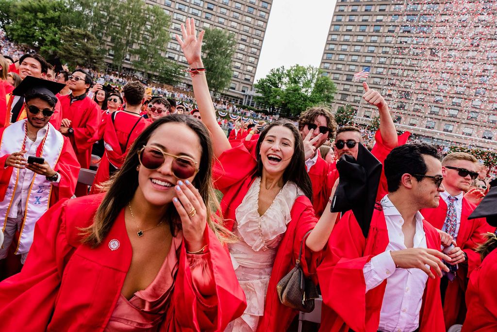 Photo of two young women cheering and smiling at the camera as red and white confetti can be seen in the background starting to fall on to the field of new graduates. The tan woman in the front pushes up her stylish aviator sunglasses and showcases her baby blue fingernails and a woman behind her cheers with arms raised in a ruffled cream dress. Other grads can be seen around them posing and smiling for the camera and celebrating amongst themselves. Everyone is wearing red gowns and either wearing or holding black caps.