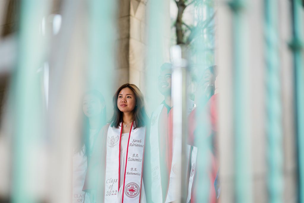 A group of students stand underneath the arches in Marsh plaza. The photo was taken behind white, iridescent streamers so the photo is blurred. A young AAPI woman at center is seen smiling, in focus.